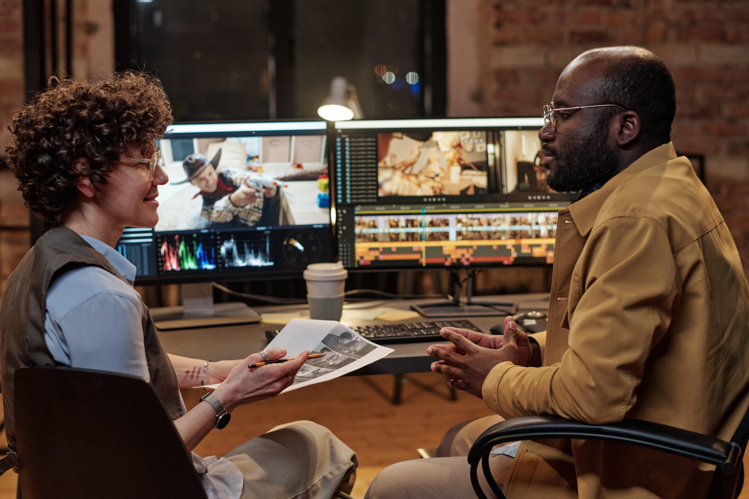 Two multiethnic colleagues discussing shooting script together while sitting opposite table with two display computers